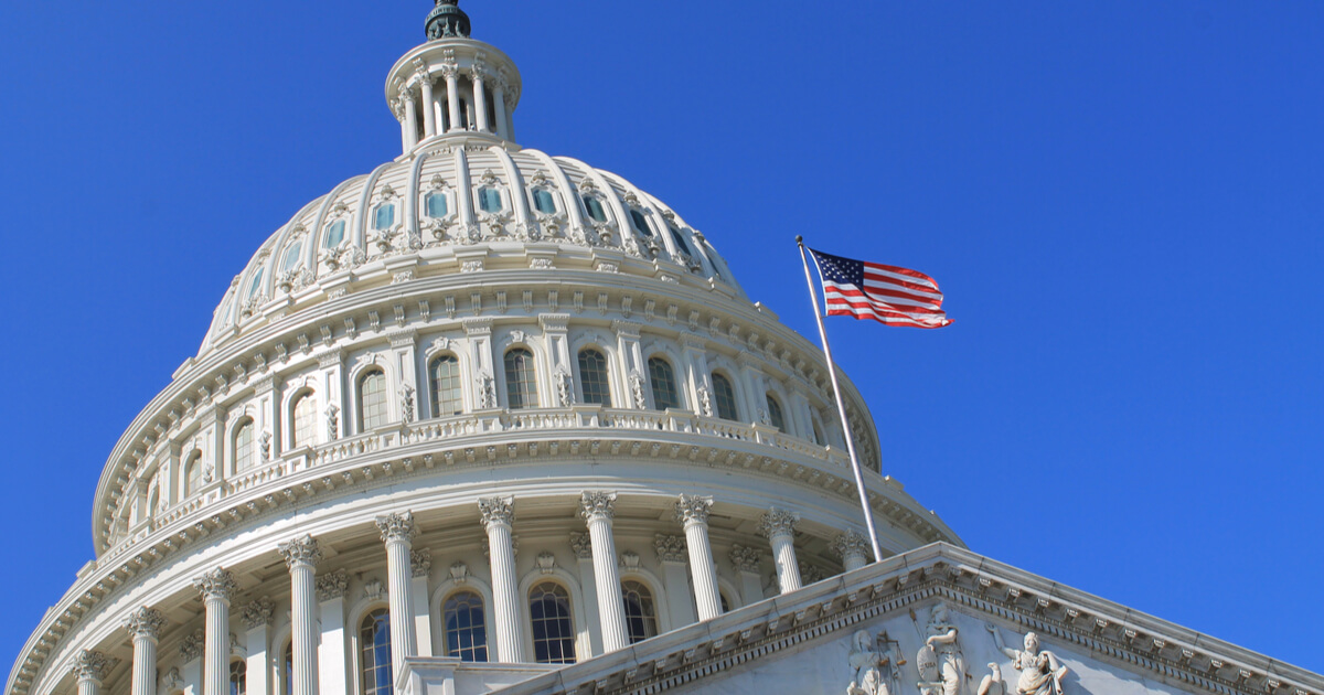 the U.S. capitol building against a clear blue sky and the U.S. flag blowing in the wind.